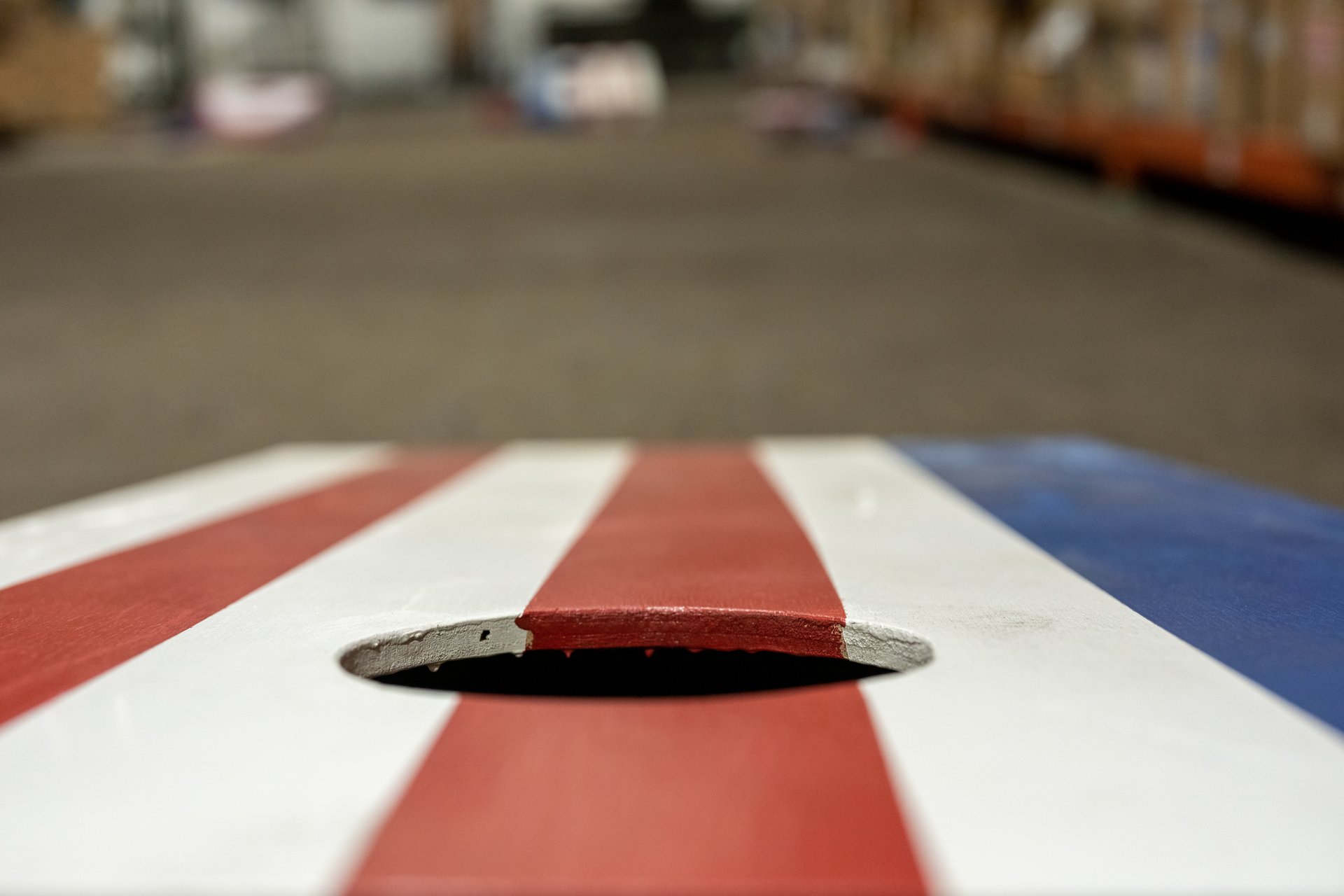 This close-up, low-angle shot features a cornhole board painted with red, white, and blue stripes, highlighting the hole in the center with a slightly worn edge, all set against a blurred background of what appears to be a warehouse or large indoor space. The mood is casual and ready for play.
