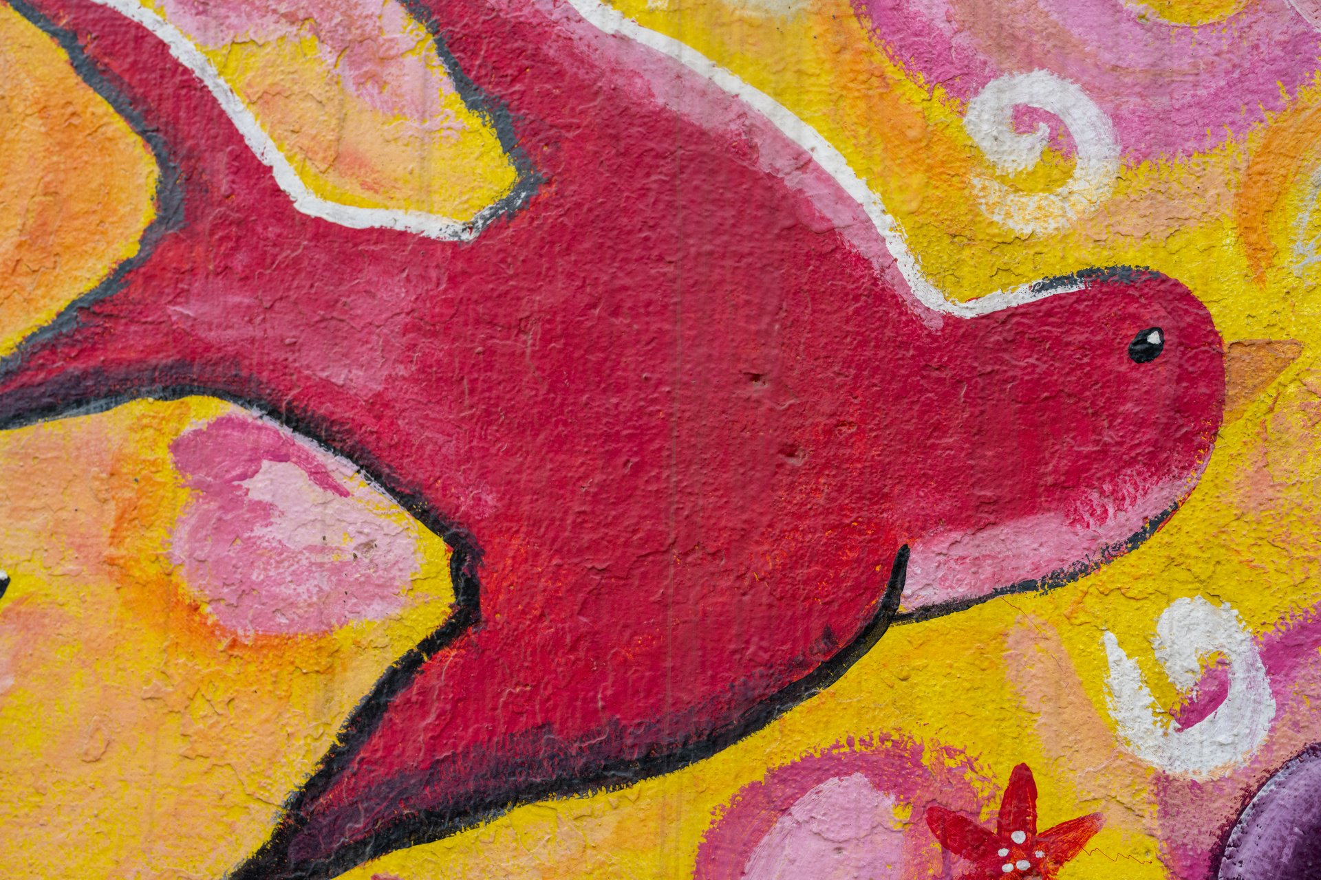 This close-up image features a vibrant red bird painted on a textured wall, surrounded by swirling patterns and floral elements in shades of yellow, pink, and white, creating a cheerful and artistic mood.