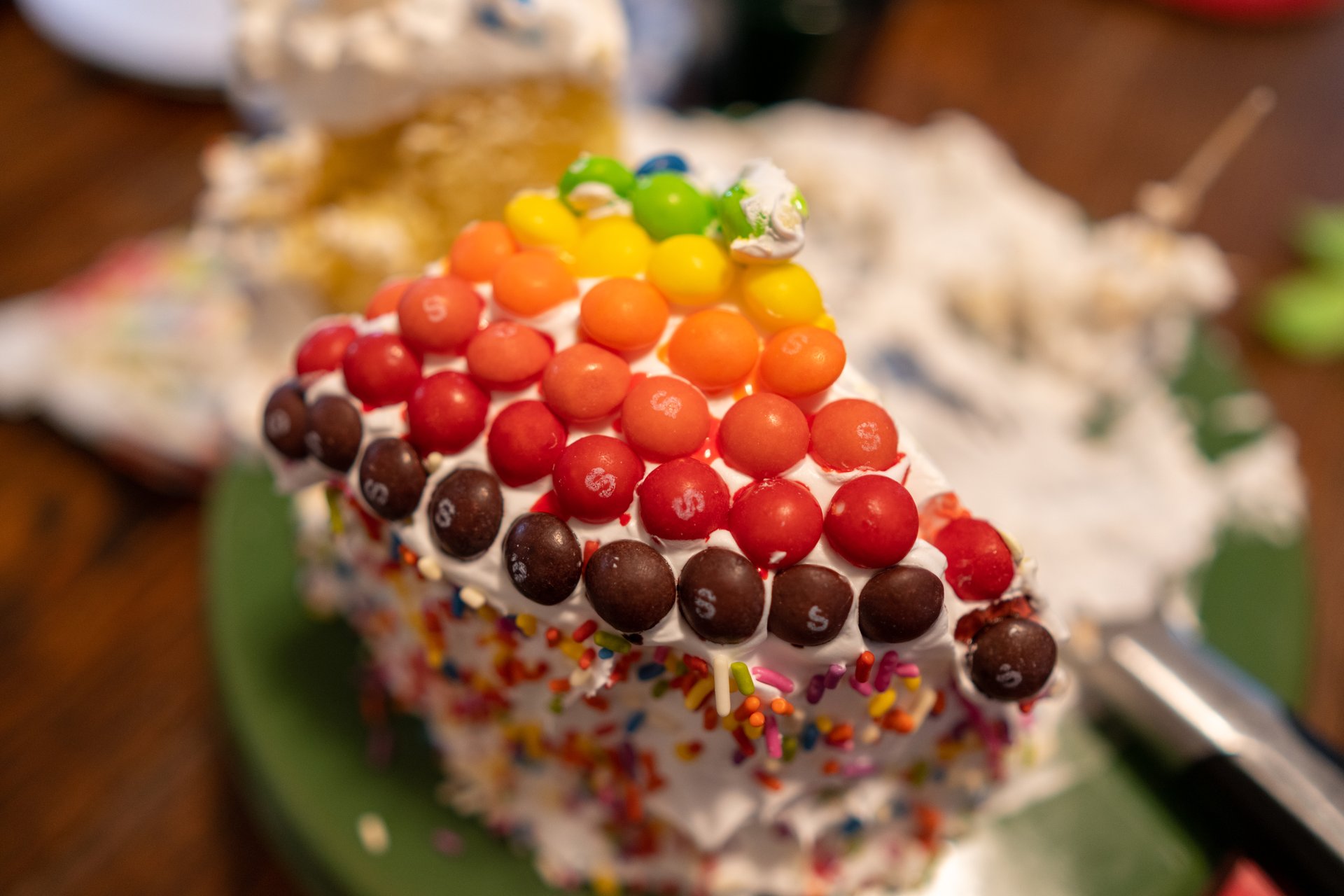 A vibrant slice of cake, covered in white frosting and colorful sprinkles, is topped with a rainbow gradient of M&M-like candies, creating a festive and playful mood.