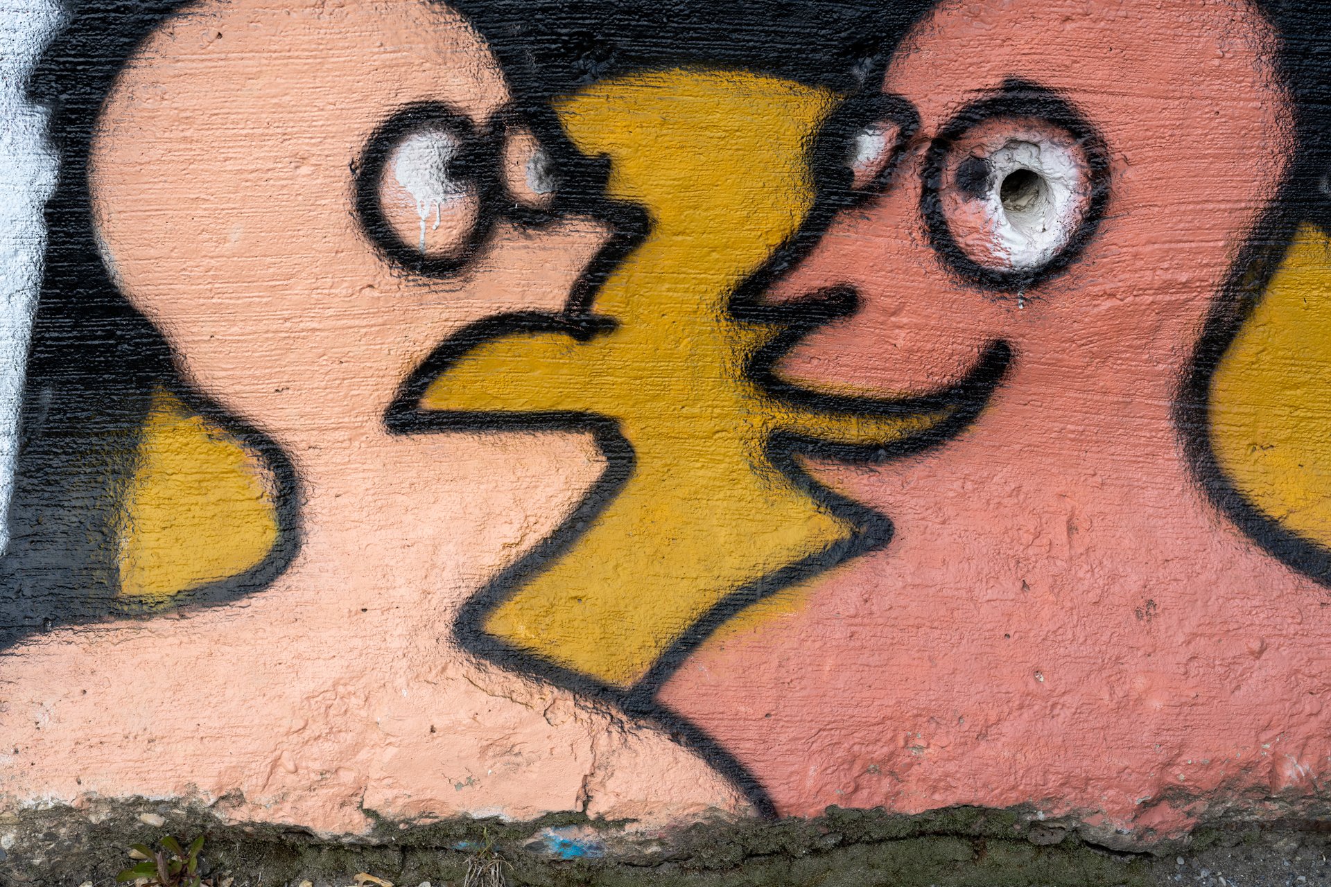 This close-up image features a vibrant graffiti artwork on a textured wall, depicting two stylized, peach-colored faces with simple black outlines and smiles, seemingly in conversation with a bright yellow, lightning bolt-like shape between them. The overall mood is whimsical and artistic, with a raw, urban aesthetic.