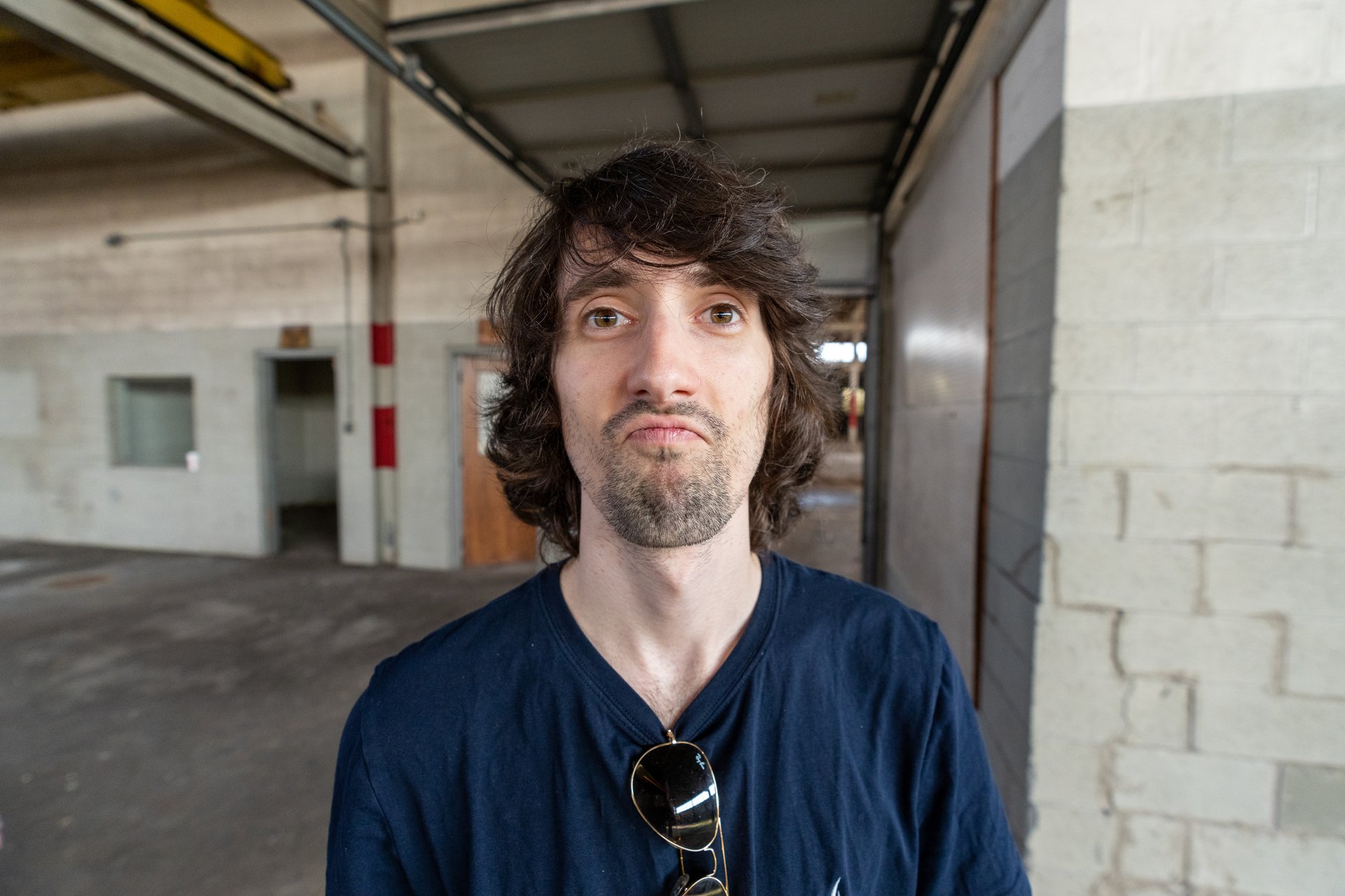A man with shaggy dark hair and a short beard stares directly at the viewer with a slightly puzzled or uncertain expression, standing in what appears to be an industrial or abandoned building with exposed beams and brick walls. He wears a dark blue t-shirt with sunglasses hanging from the collar, conveying a casual and somewhat contemplative mood.