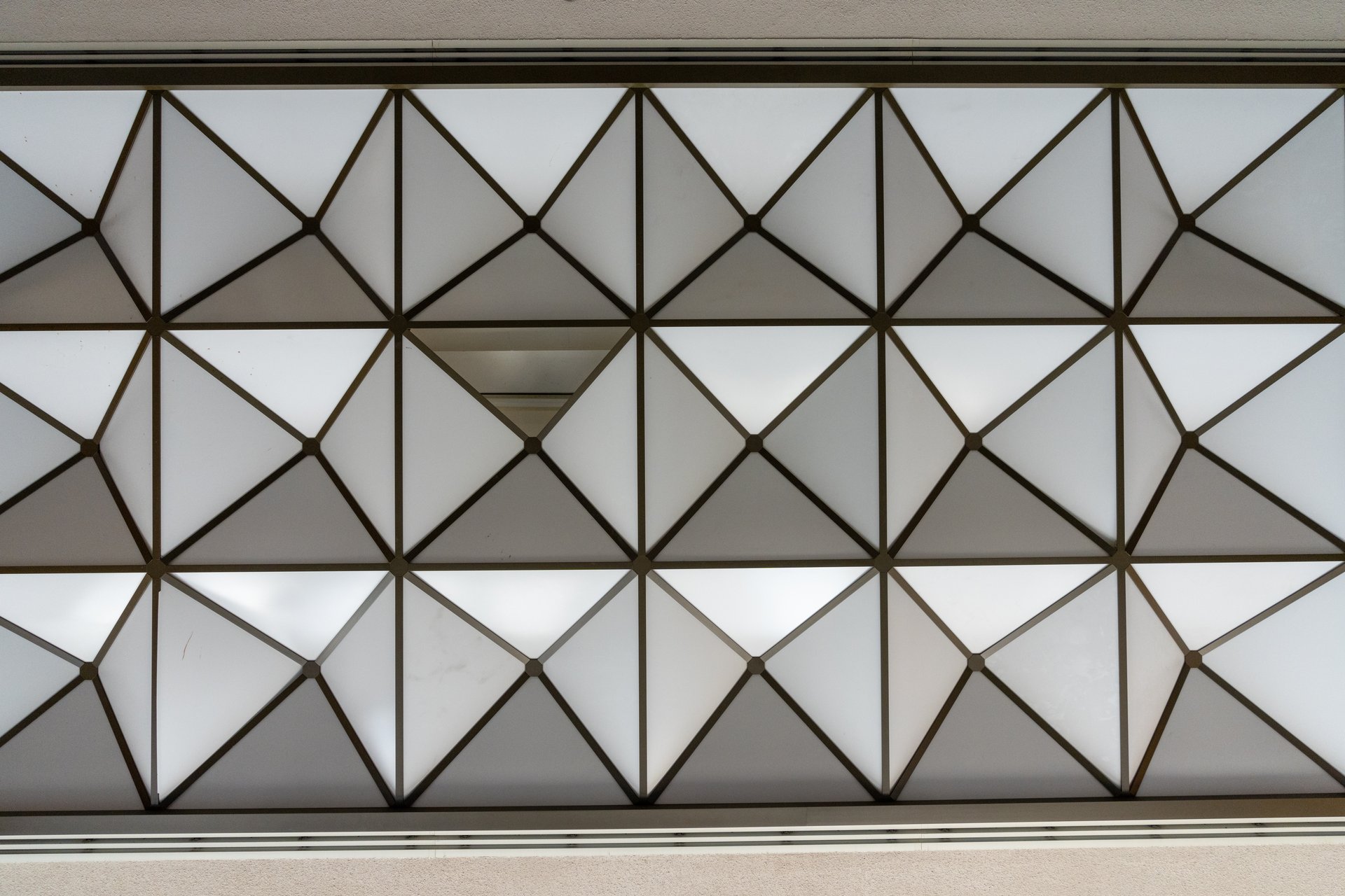 This image displays a modern ceiling design featuring a repeating pattern of triangles and squares in white translucent panels framed by dark lines, creating a structured and slightly abstract mood.