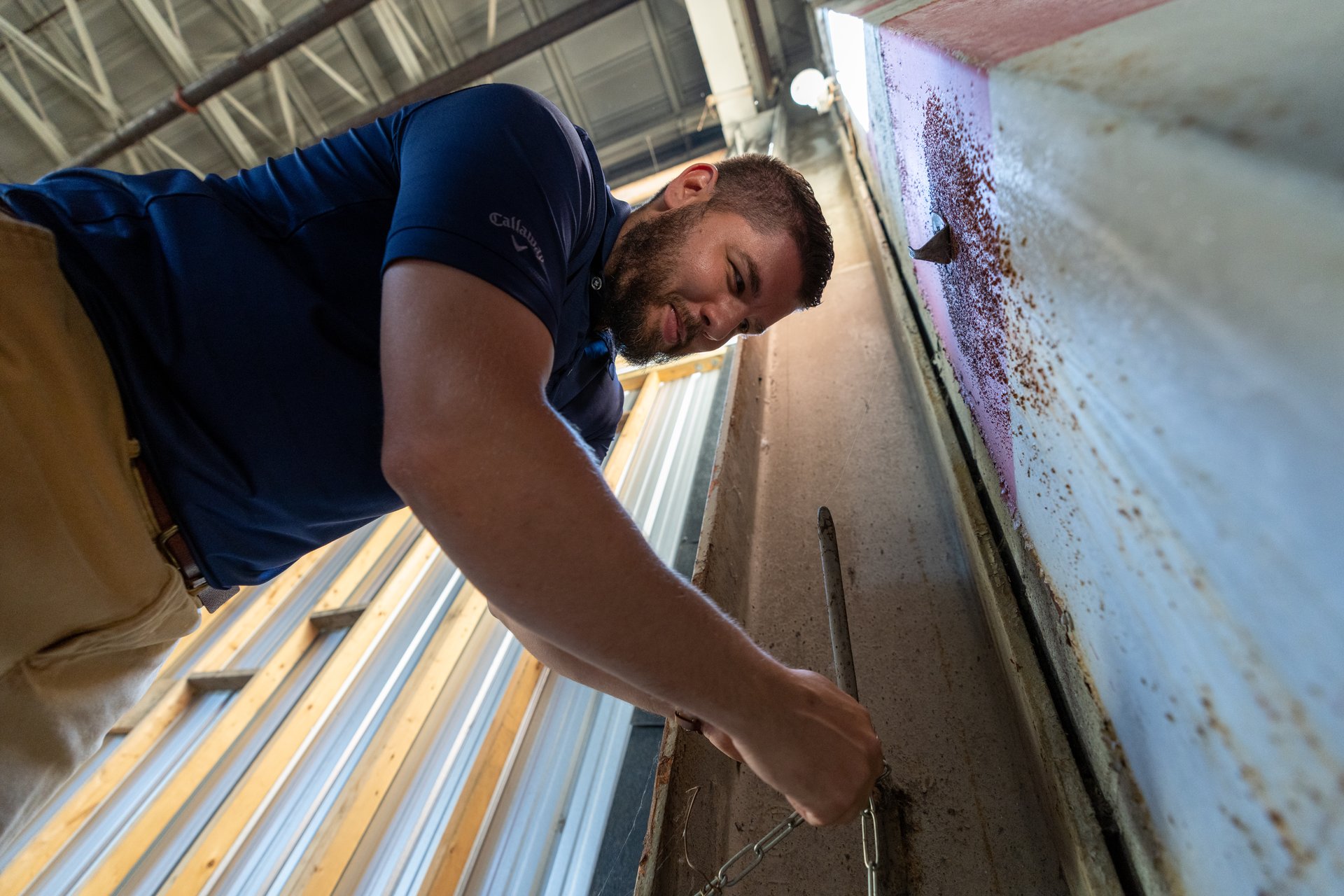 A man with a beard, wearing a blue polo shirt and khaki pants, is intently focused on working with chains attached to a metal surface, possibly in an industrial or workshop setting. The low-angle shot and his concentrated expression suggest a task requiring precision and effort.