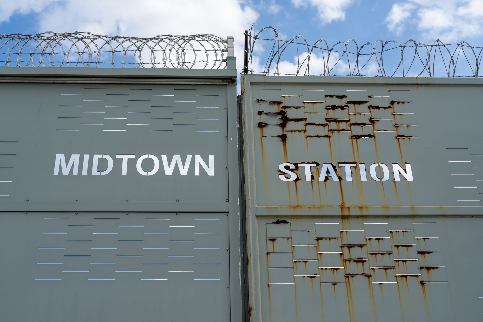 This image shows a large, grey metal gate with the words "MIDTOWN STATION" stenciled in white. The gate is topped with barbed wire, and the right panel shows significant rust and wear, conveying a sense of security and perhaps neglect under a bright, cloudy sky.