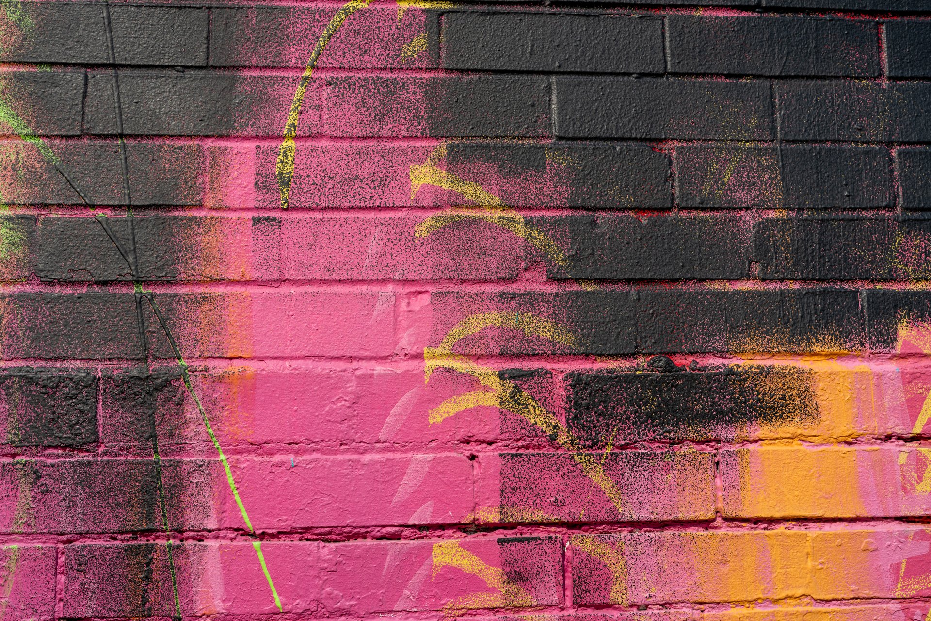 This close-up image features a brick wall heavily adorned with vibrant graffiti in shades of pink, yellow, and black, creating an energetic and urban mood. The layered paint and visible brick texture suggest a dynamic and well-used public space.