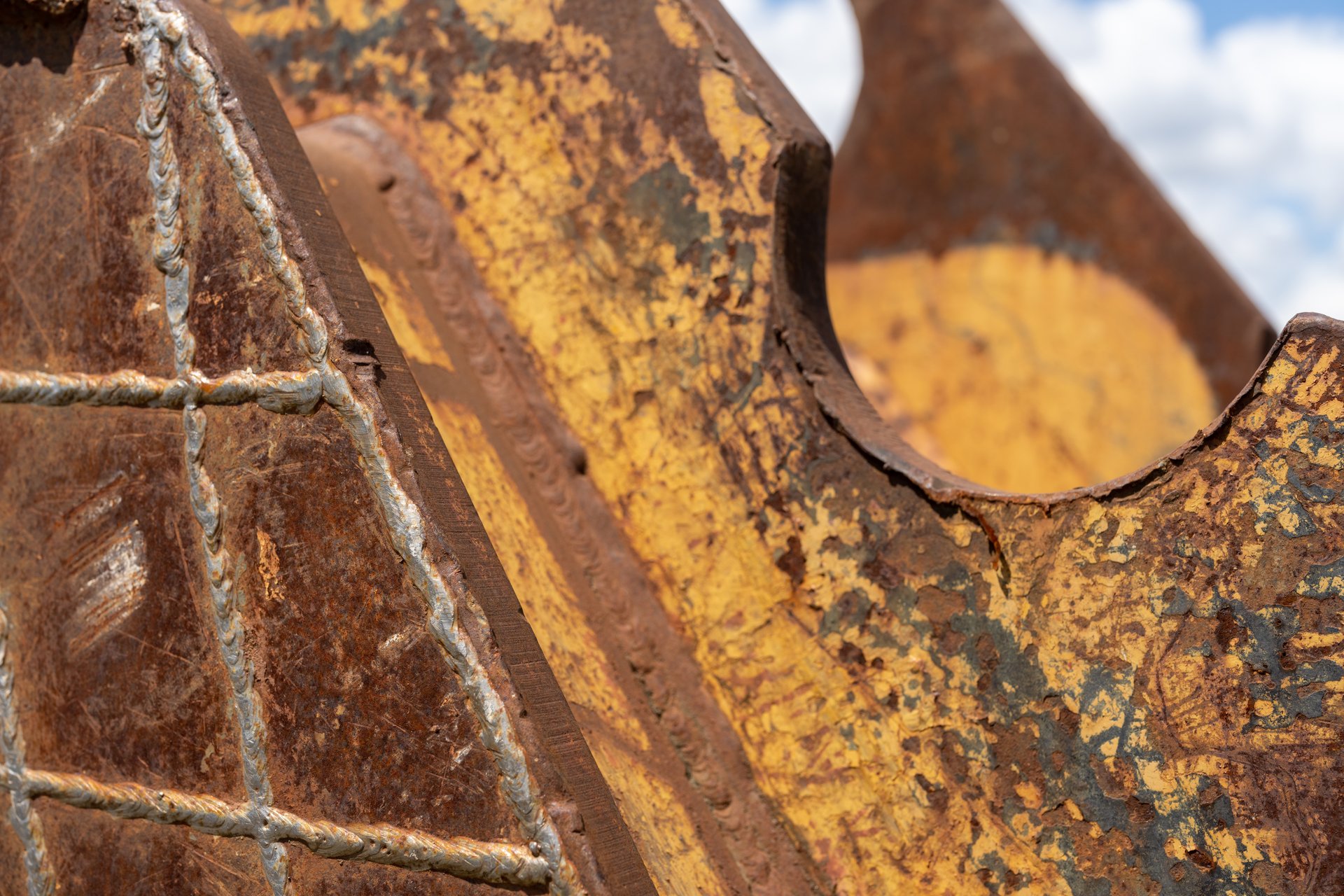 This close-up shot reveals the heavily rusted and weathered surface of what appears to be part of heavy machinery or an industrial structure, showcasing a rich palette of browns, oranges, and faded yellows against a blurred background of a cloudy sky. The intricate textures of corrosion and welding seams create a sense of age and hard use.