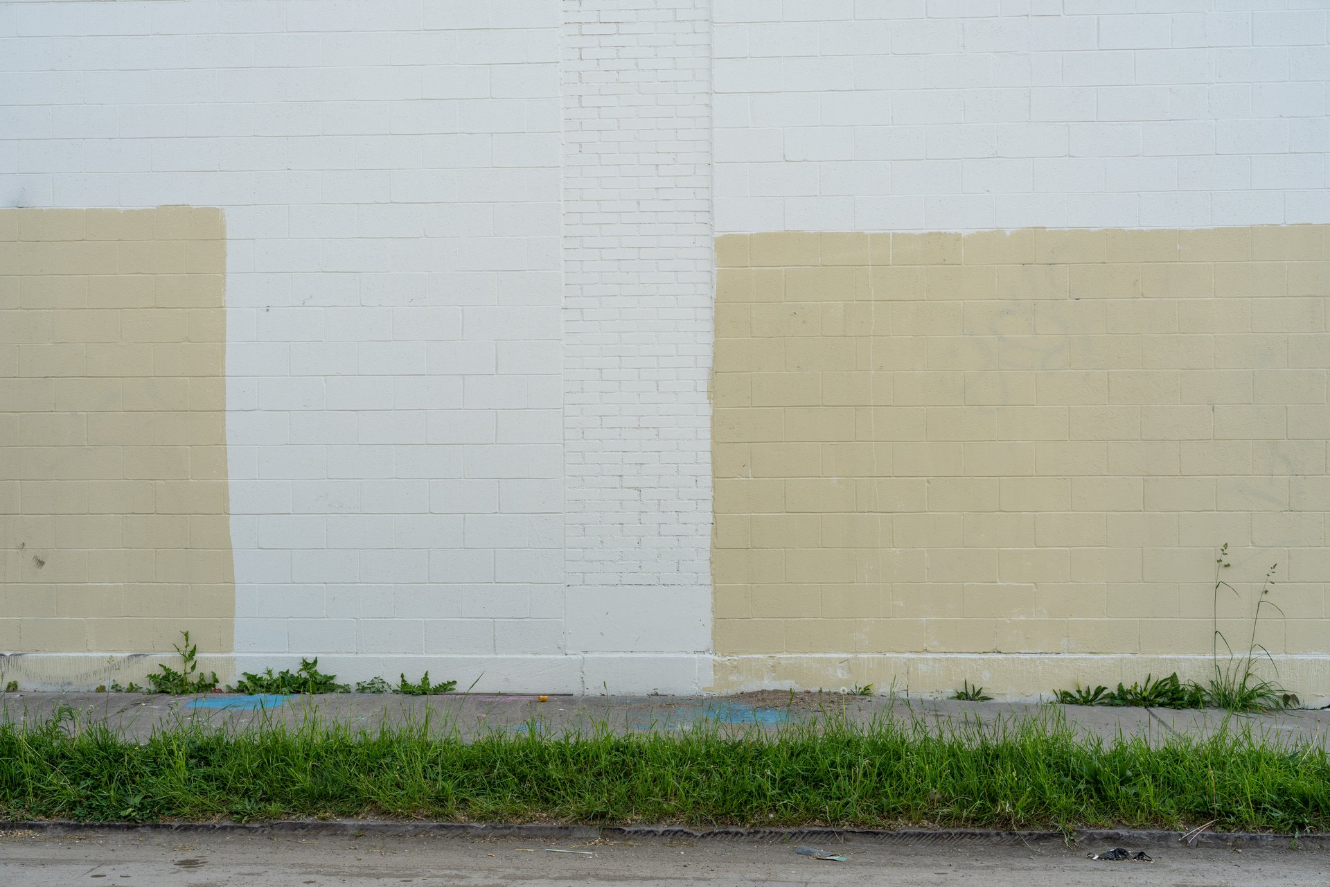 This image captures a white brick wall with two rectangular sections painted in a light tan color, suggesting an unfinished or abandoned painting project. A strip of green grass and weeds grows along the bottom, separating the wall from a narrow concrete sidewalk, creating a somewhat neglected urban mood.