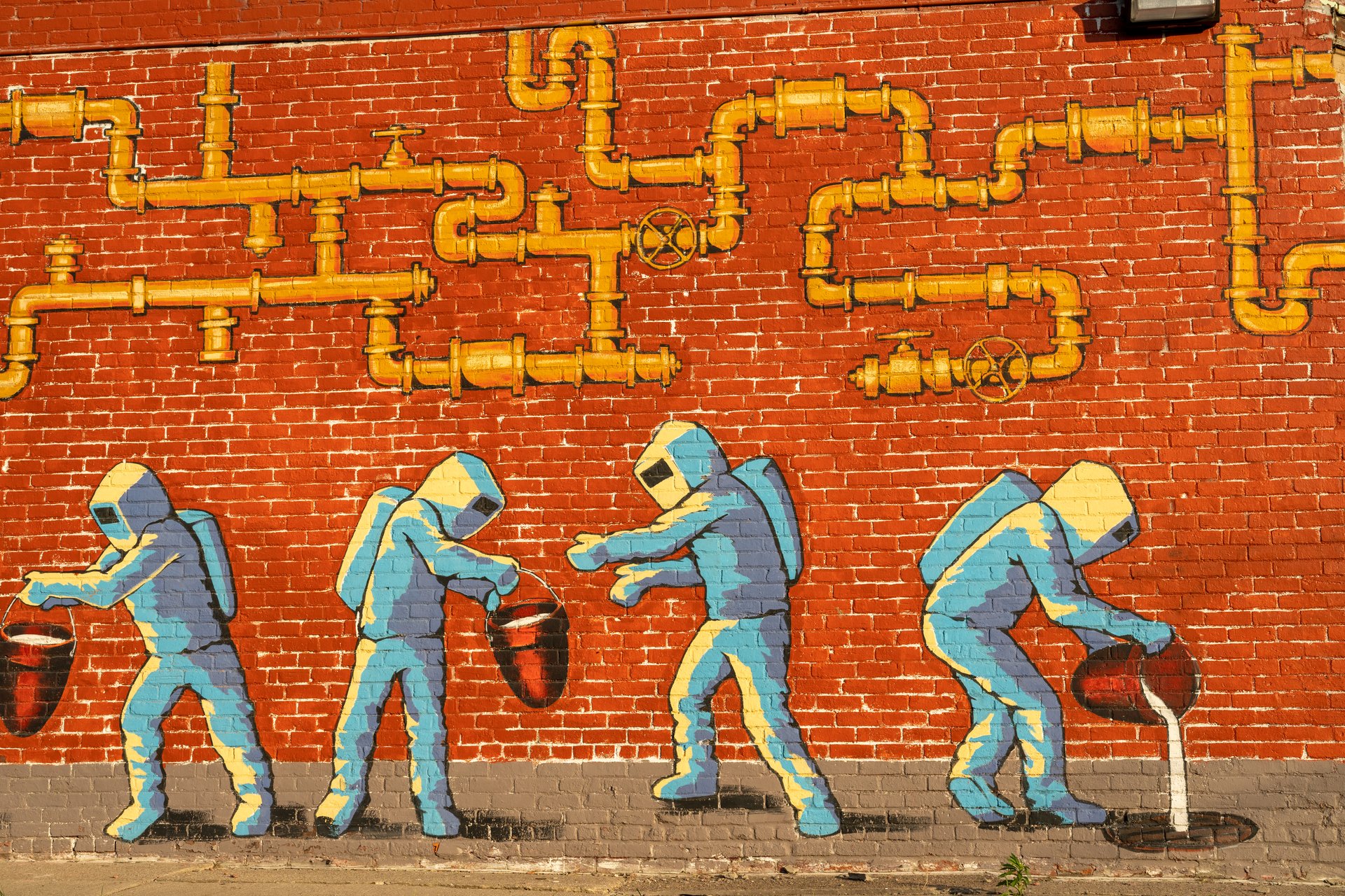 This vibrant mural on a red brick wall depicts four figures in light blue industrial suits, engaging in tasks involving buckets, beneath a complex network of yellow pipes and valves. The bright colors and clear lines create a striking and somewhat whimsical portrayal of labor.
